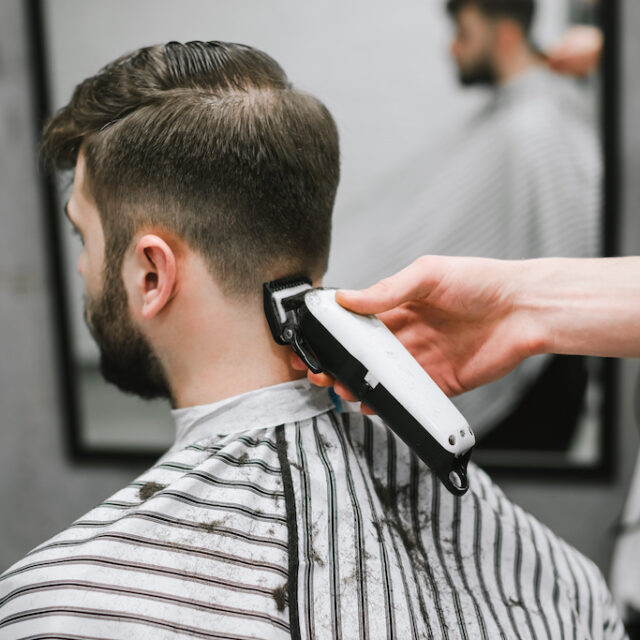 Professional male hairdresser does hairstyle to bearded man in barber shop, back view, barber's hand with clipper. Barber clipping client in men's hairdresser, closeup photo